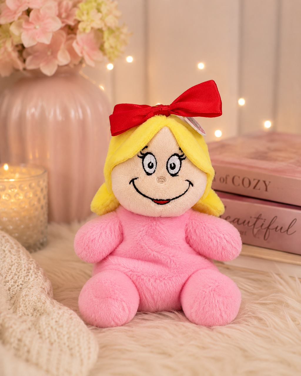 Cindy Lou Who palm pal plush toy sitting on a fluffy cream rug beside a candle and pink books, red bow and yellow pigtails visible against a soft pink and floral background.