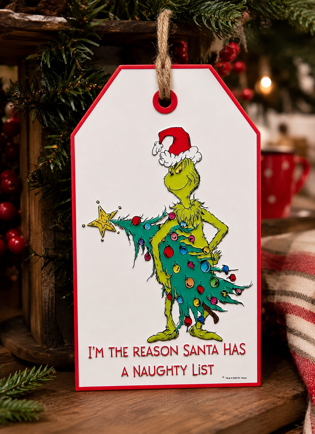 Grinch naughty list hanging sign displayed on a wooden crate surrounded by pine branches and berries, red mug and plaid blanket in background, Grinch wrapped in Christmas tree with "I'm the reason Santa has a naughty list" text.