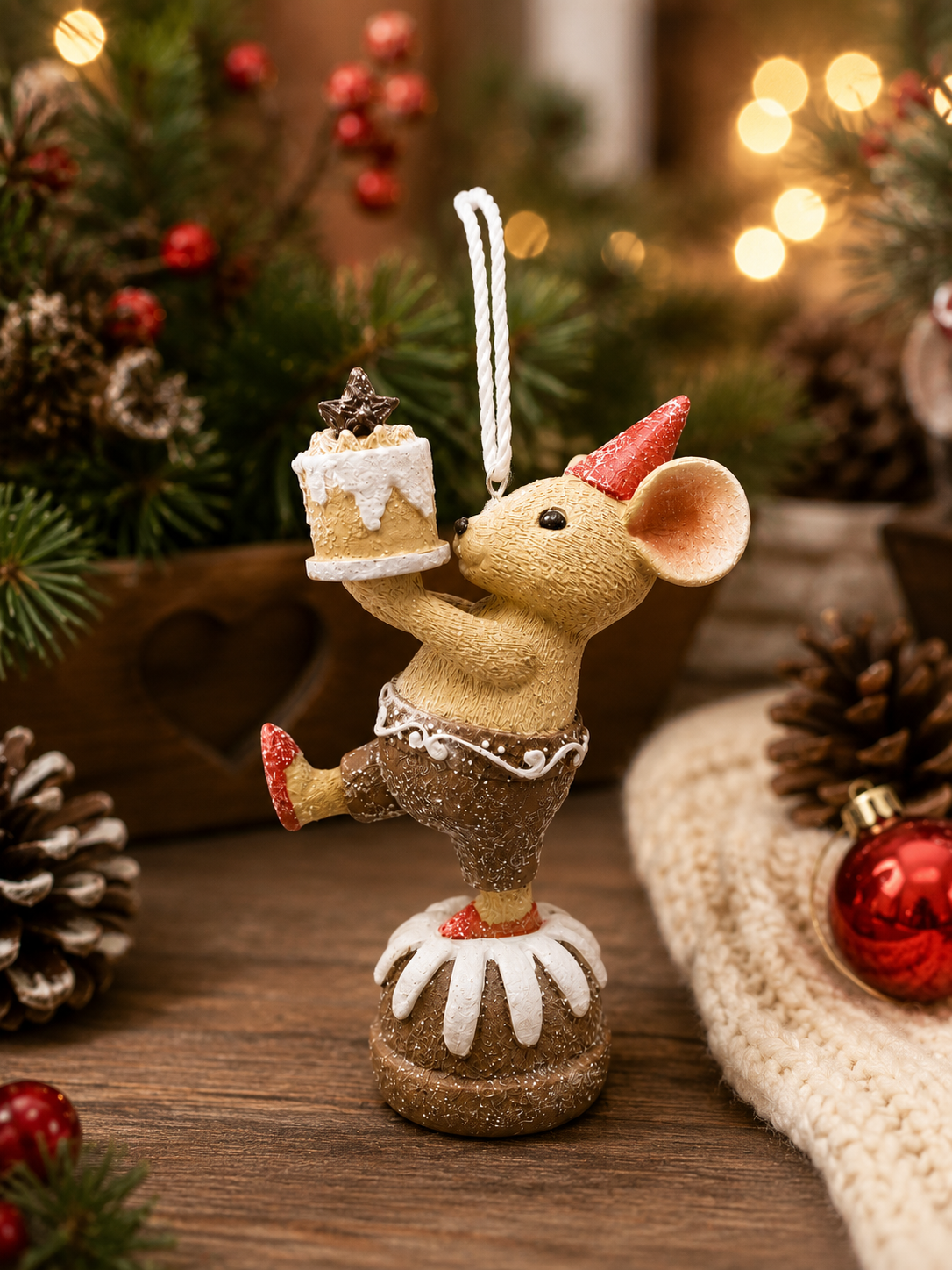Party cake mouse hanging ornament displayed among pine branches and pinecones, mouse in red party hat holding an iced cake while balanced on a Christmas pudding, red bauble in foreground.