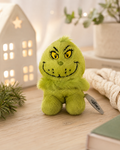 Palm Grinch plush toy sitting on a wooden table beside a glowing house ornament and pine sprigs, showing embroidered grinning face and yellow eyes