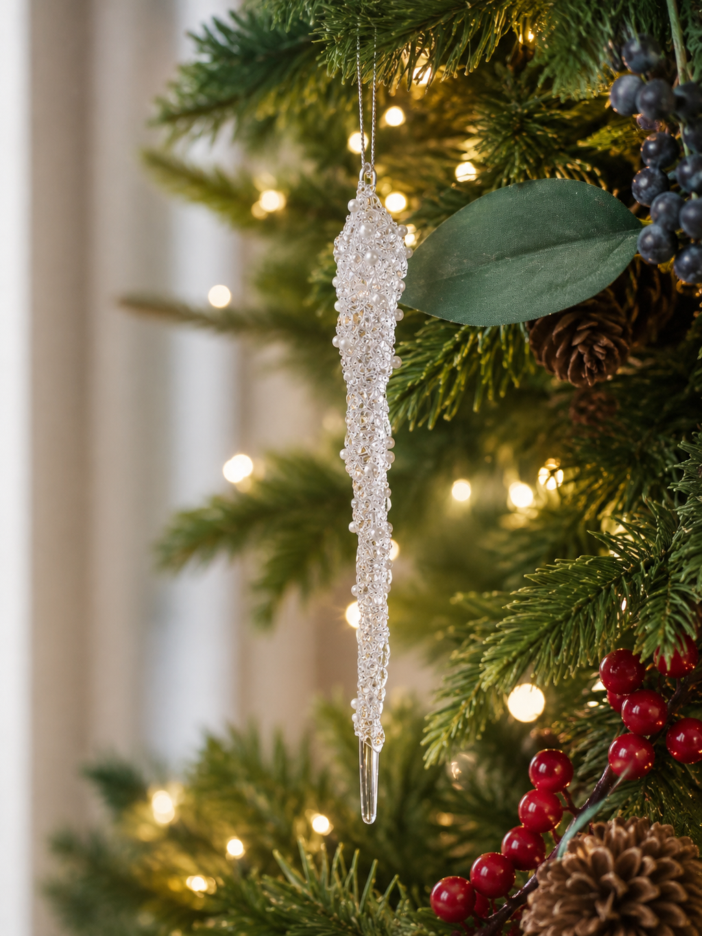 Pearl and crystal encrusted icicle Christmas ornament hanging on a lit Christmas tree with magnolia leaves, blue berries and red berry clusters, glowing softly in the warm tree lights
