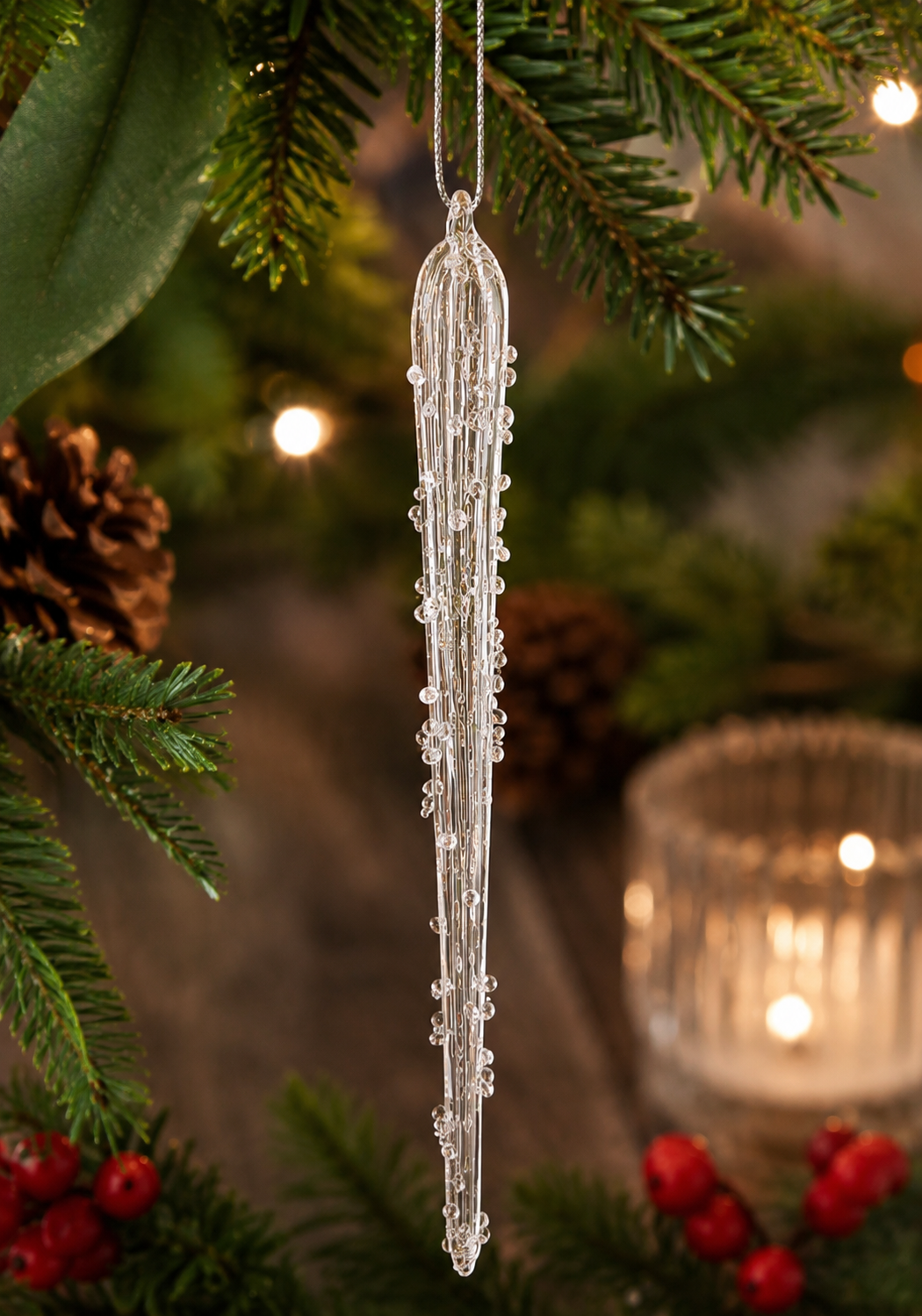 Clear acrylic crystal droplet icicle Christmas ornament hanging on a lit Christmas tree with pine cones and magnolia leaves, glowing in warm candlelight with a ribbed strand texture and scattered droplet beads