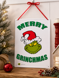Grinch "Merry Grinchmas" tin sign hanging on a wall beside a frosted Christmas tree, styled with a red bauble, burlap pumpkin and pinecones on a wooden shelf