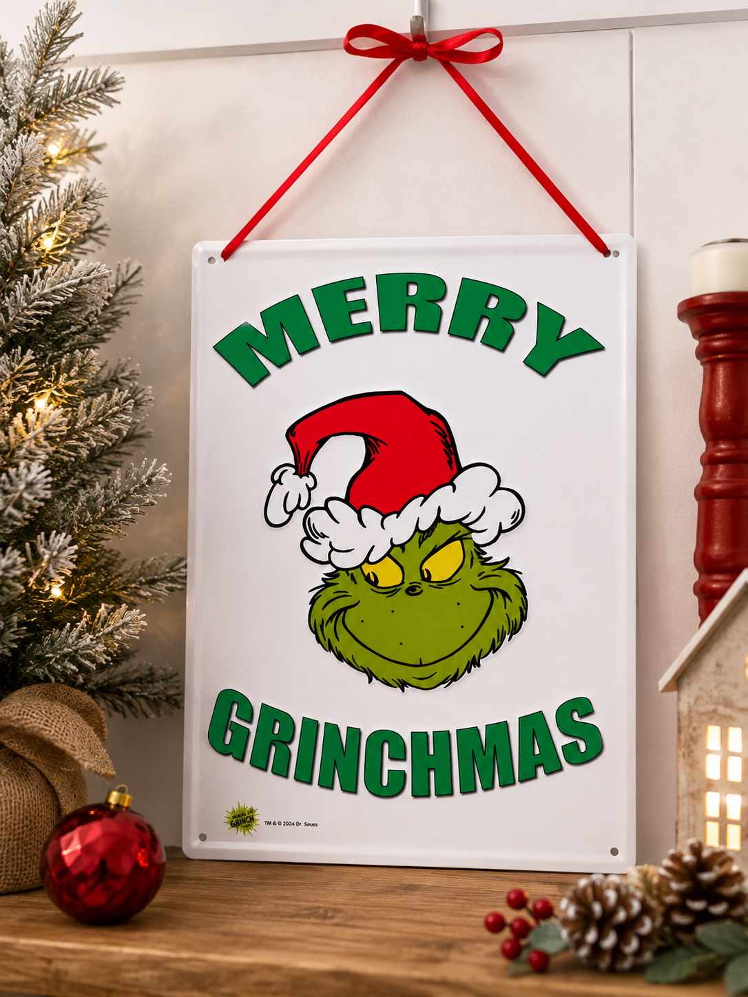 Grinch "Merry Grinchmas" tin sign hanging on a wall beside a frosted Christmas tree, styled with a red bauble, burlap pumpkin and pinecones on a wooden shelf
