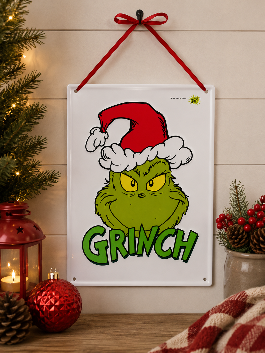 Grinch face tin sign hanging on a wall beside a lit Christmas tree, styled with a red lantern, candle, bauble and pinecones on a wooden shelf