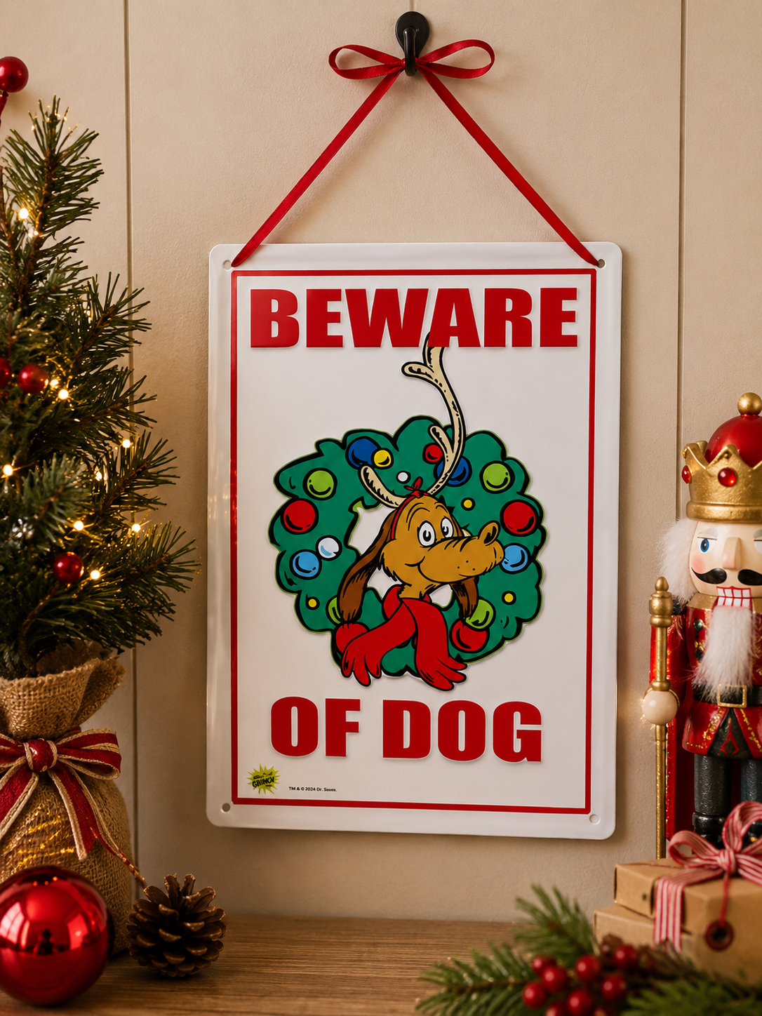 Grinch "Beware of Dog" tin sign featuring Max the dog in a wreath, hanging on a wall beside a lit Christmas tree with baubles, pinecones and wrapped gifts