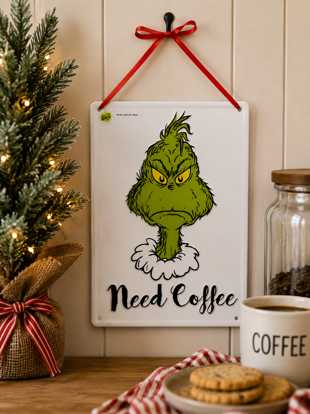 Grinch "Need Coffee" tin sign hanging on a wall near a coffee jar, mug and biscuits, beside a lit Christmas tree