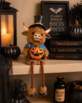 Highland cow Halloween figurine in top hat and blue scarf holding glowing jack-o-lantern, seated on wooden shelf beside "Hocus Pocus" sign, spell books and witches brew bottle