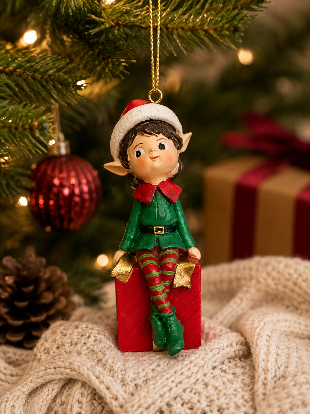 Elf boy on gift box ornament hanging from a Christmas tree branch, red bauble, pinecone and wrapped gift in the background
