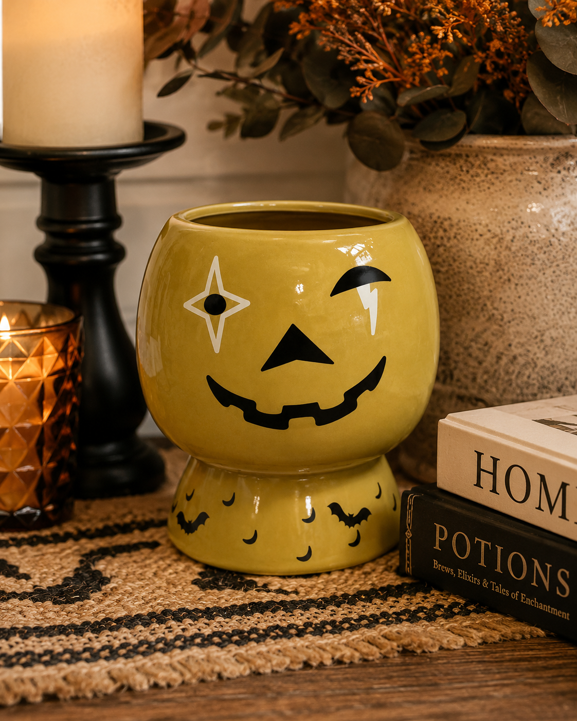 Jack-o'-lantern ceramic plant pot on a woven table runner beside a candle, lantern and autumn foliage, with stacked books