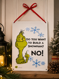 Grinch "Do You Want to Build a Snowman? NO!" hanging sign displayed on a wall beside a lit candle, lantern and snow-dusted mini tree