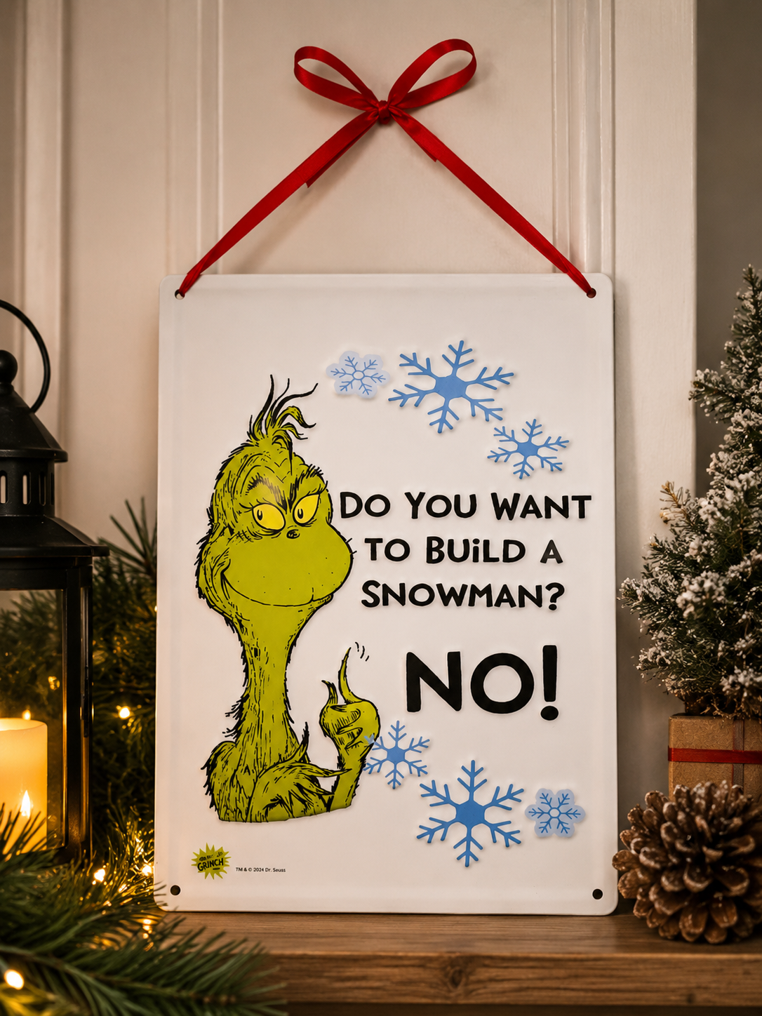 Grinch "Do You Want to Build a Snowman? NO!" hanging sign displayed on a wall beside a lit candle, lantern and snow-dusted mini tree
