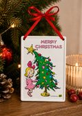 Cindy-Lou Who tin sign hanging near a Christmas tree, red bauble and glass candle holder, with pinecones nearby