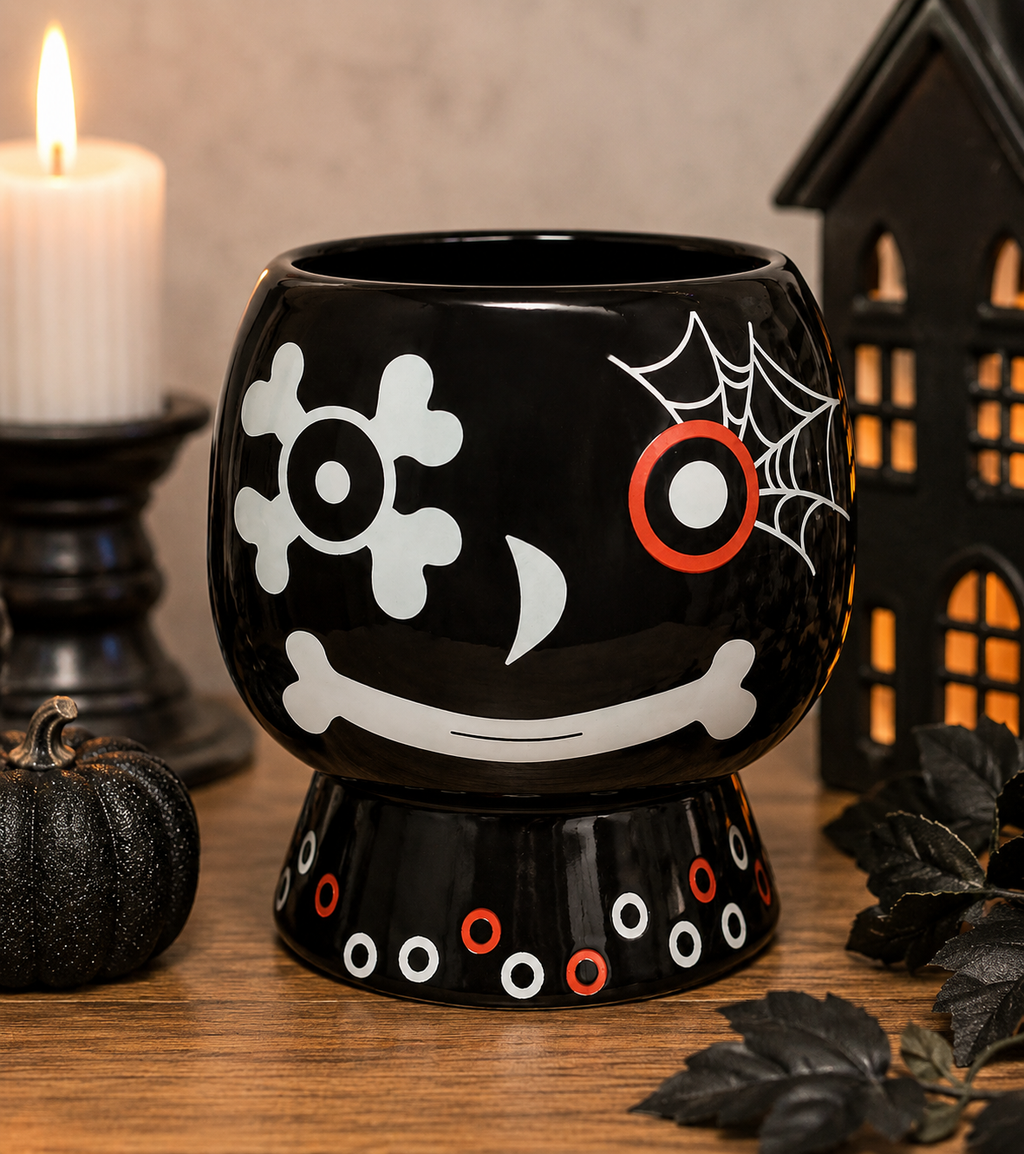 Skull and web ceramic plant pot on a wooden table beside a lit candle, black pumpkin and haunted house lantern