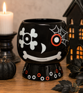 Skull and web ceramic plant pot on a wooden table beside a lit candle, black pumpkin and haunted house lantern