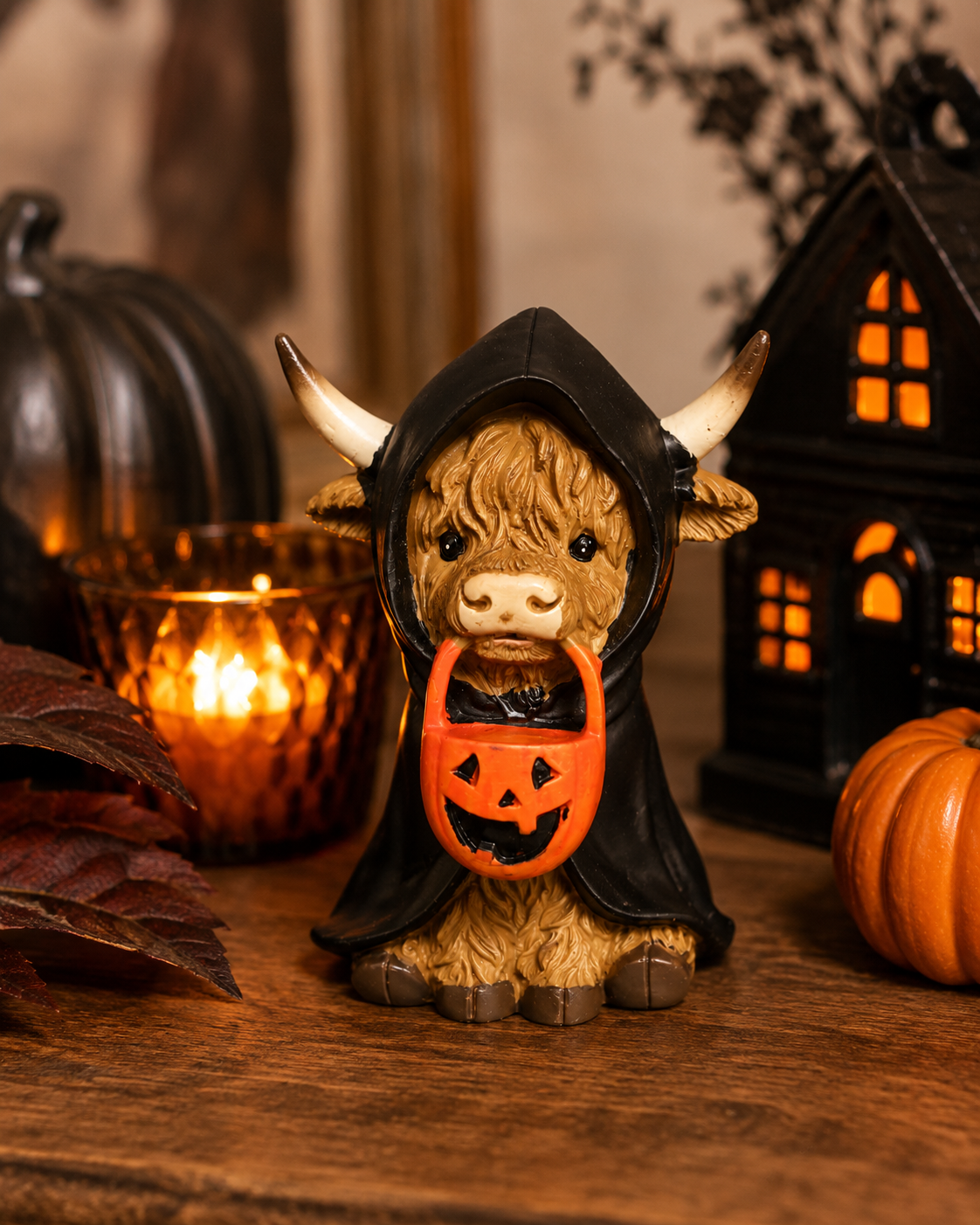 Highland cow figurine in a black reaper cloak holding a jack-o'-lantern bucket, displayed on a wooden table beside candles, a pumpkin and a haunted house lantern