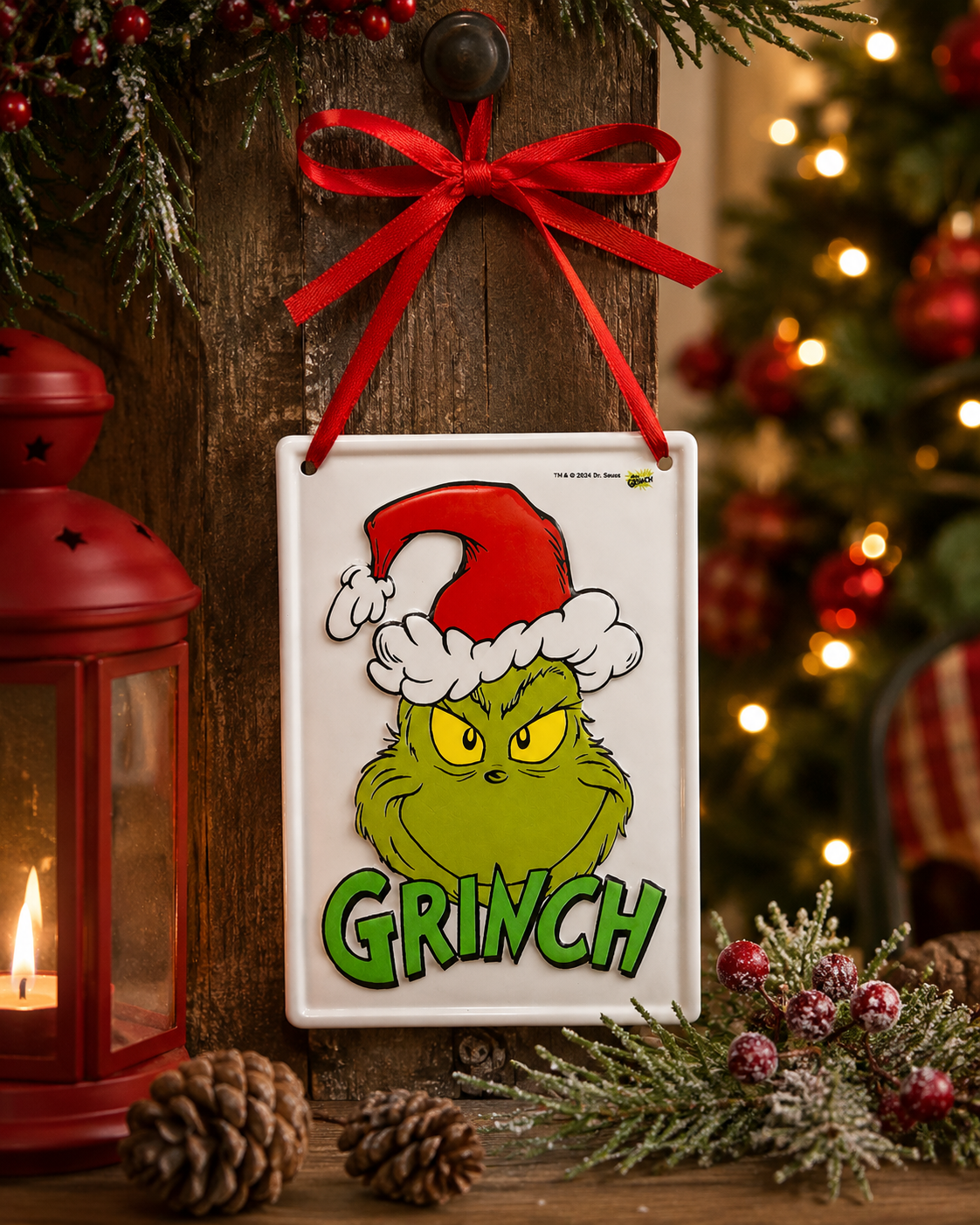 Grinch tin ornament hanging on a rustic wooden post beside a Christmas tree, red lantern and pinecones, red ribbon bow at top