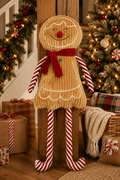 Gingerbread girl plush decoration leaning against a wooden staircase post, surrounded by wrapped gifts and a Christmas tree with lights