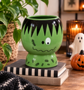 Frankenstein ceramic plant pot on a wooden table surrounded by pumpkins, black lantern and Halloween decor
Image