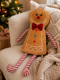 Gingerbread man plush decoration seated on a cream armchair beside a knitted throw and wrapped Christmas gifts, tree in background