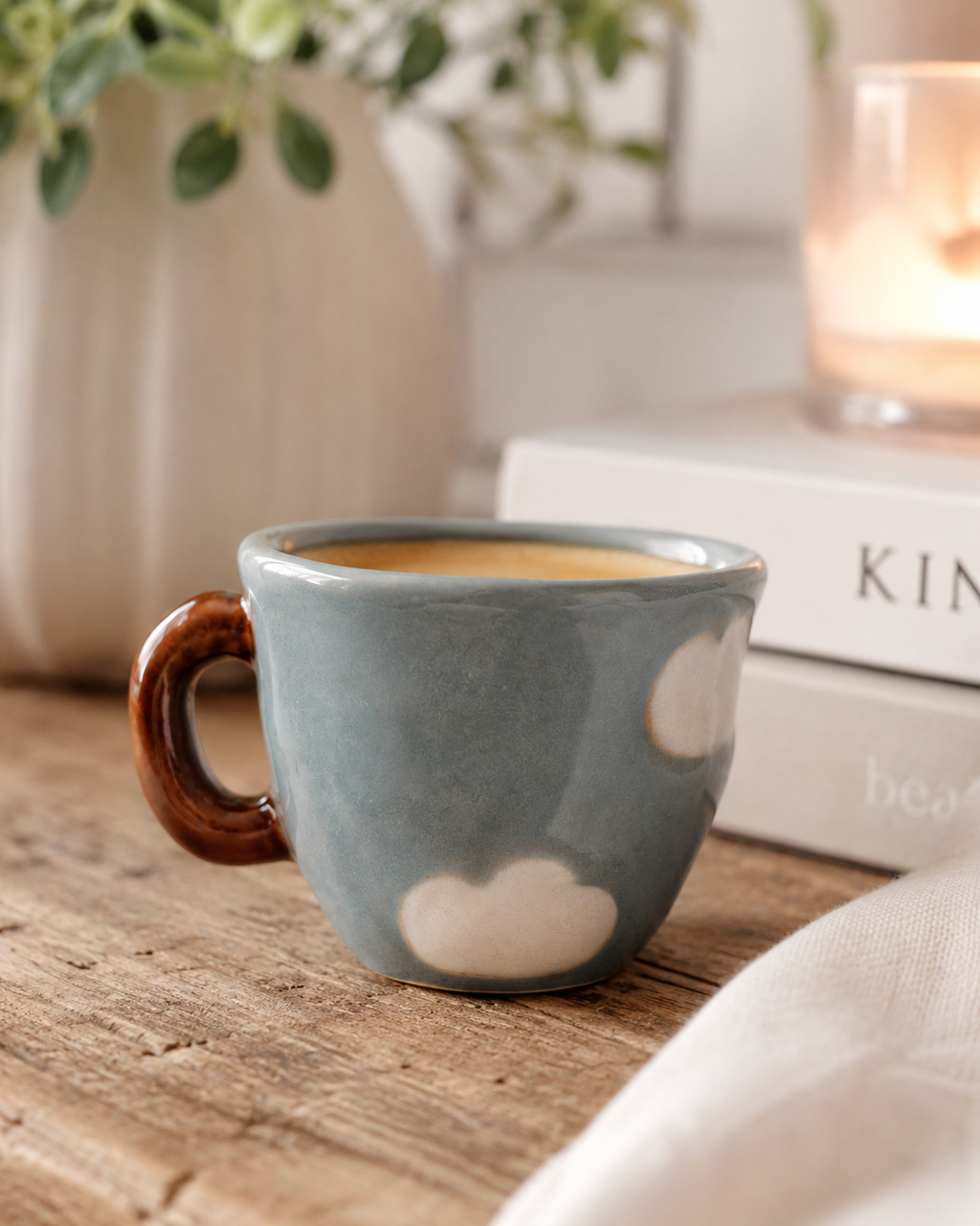 Nini Cup Clouds kids' mug filled with a warm drink on a rustic wooden table beside stacked books and a lit candle, styled with greenery in a cosy home setting