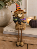 Highland cow witch figurine with black cat sitting on stacked books beside a vase and pumpkin, dangly legs hanging over the shelf edge