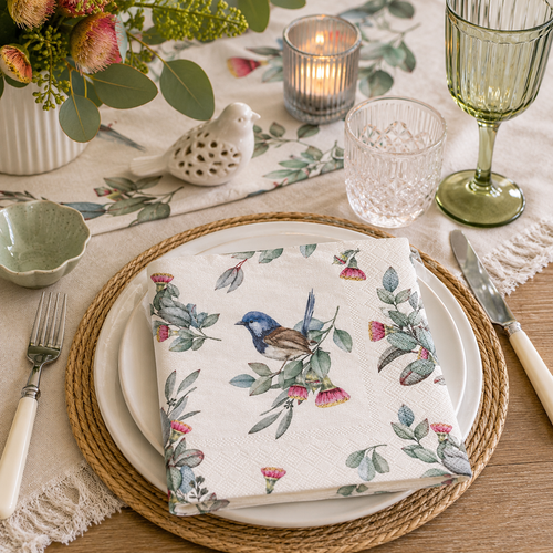 Blue wren and gum blossom paper napkin folded on layered white plates with a ceramic bird ornament, eucalyptus greenery and green glassware on a native-themed table setting