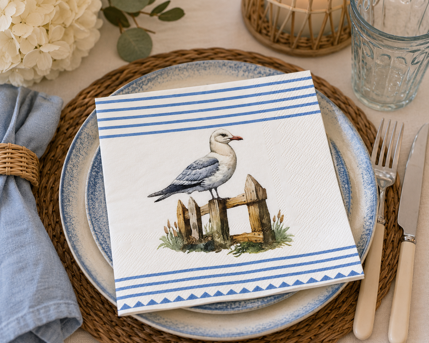 Blue and white seagull paper napkin styled on a blue-rimmed stoneware plate, with hydrangeas, a woven candle holder and cutlery on a coastal-style table setting