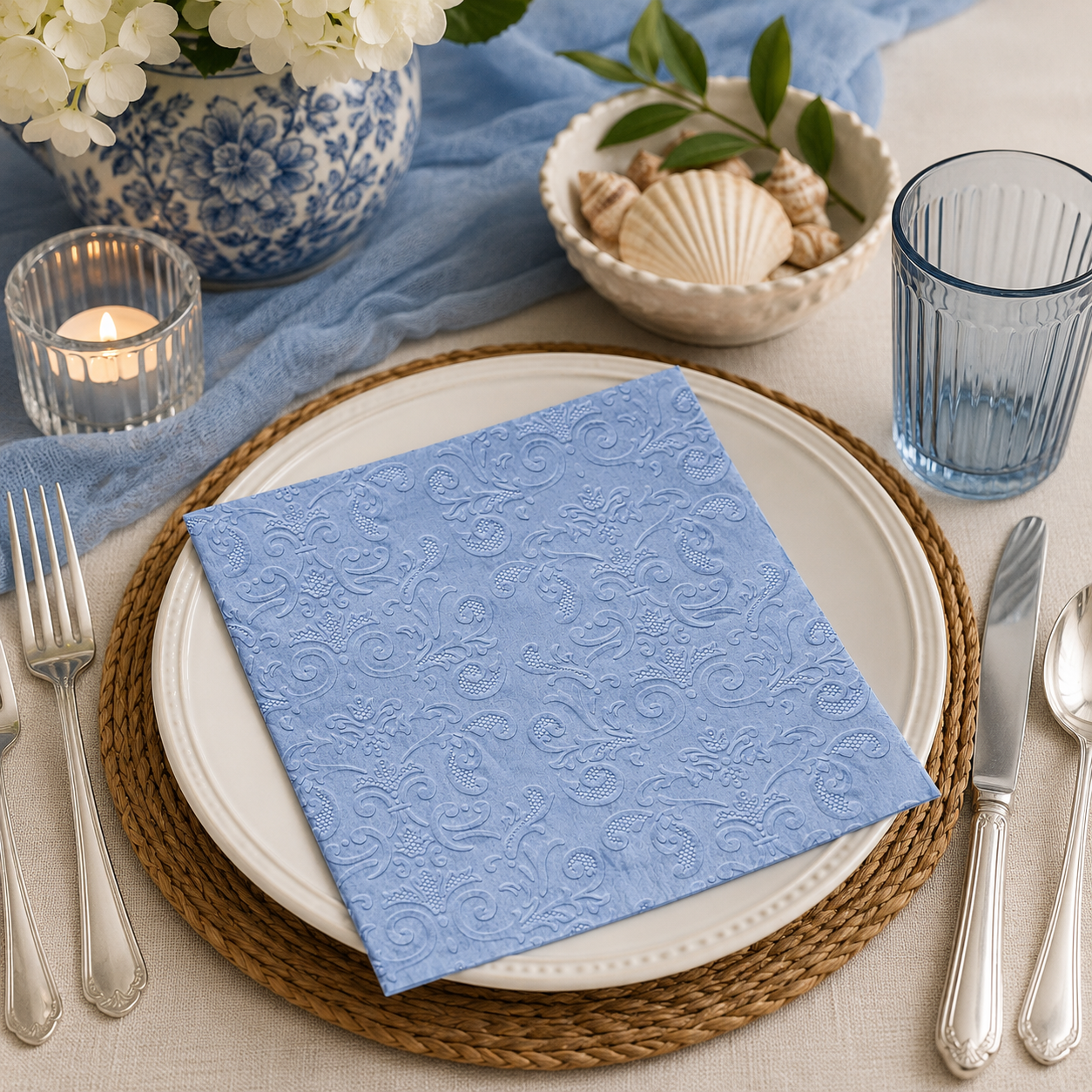 Embossed dusty blue damask paper napkin folded on a stoneware plate with a lit candle, seashells and blue glassware on a coastal table setting