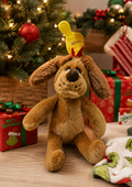 Max Chubb's plush dog sitting under a decorated Christmas tree, wearing his red and yellow antler, surrounded by Merry Grinchmas gift boxes and a striped stocking