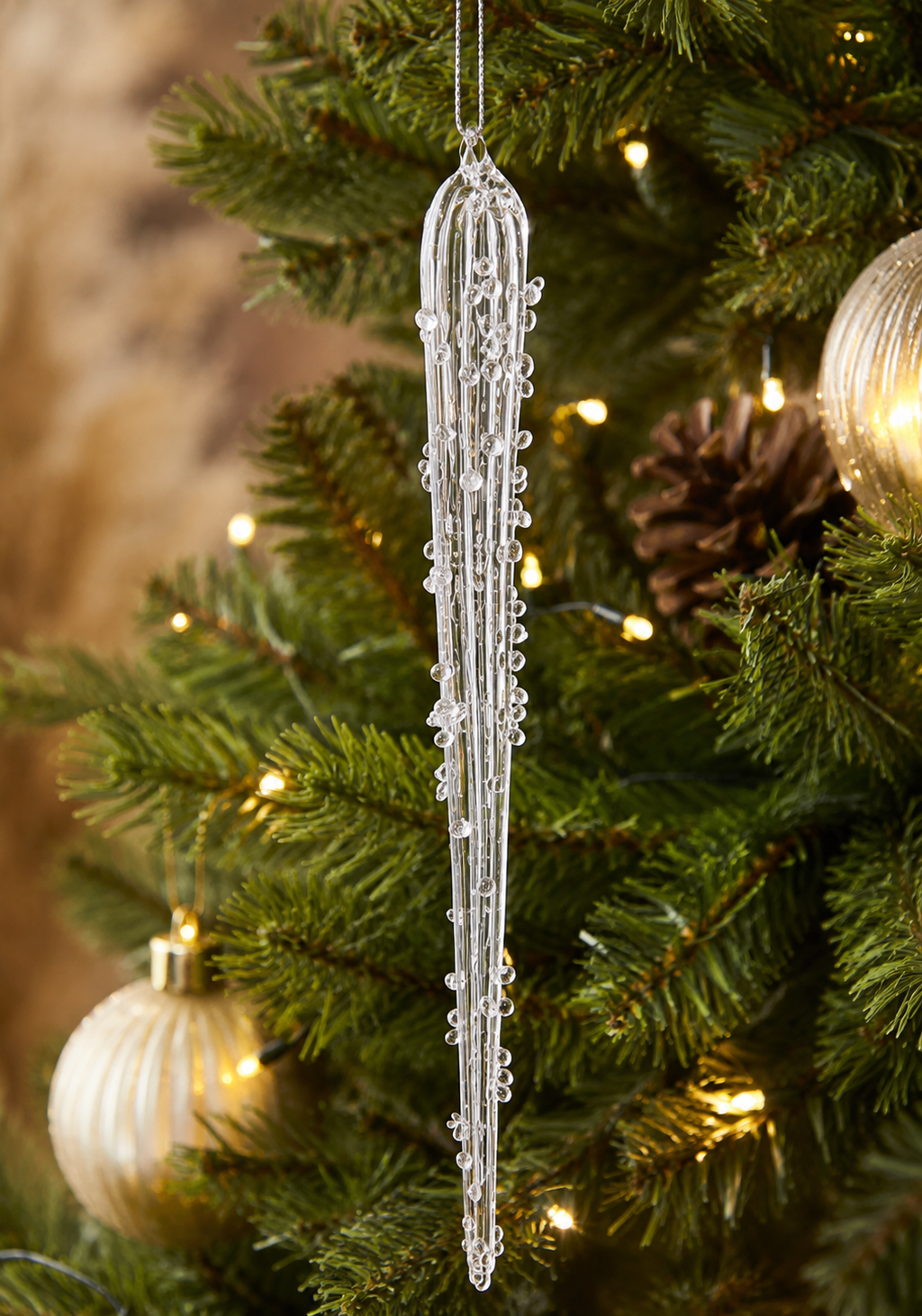 Encrusted glass icicle ornament hanging on a Christmas tree branch near a pinecone and gold bauble