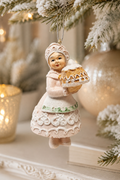 Pastel Mrs Claus pie ornament hanging on a flocked Christmas tree branch, with candlelight and a gold bauble blurred in the background