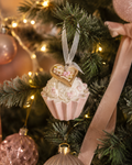 Pink and white cupcake ornament with a gold heart charm hanging on a Christmas tree branch among pink baubles and a velvet bow