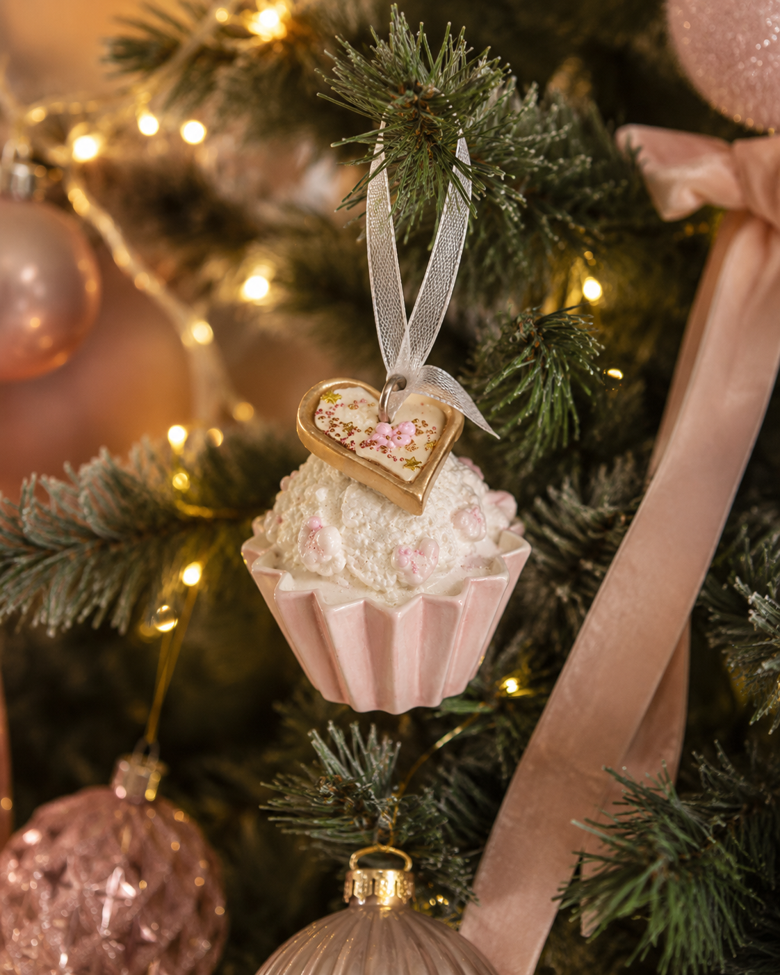 Pink and white cupcake ornament with a gold heart charm hanging on a Christmas tree branch among pink baubles and a velvet bow