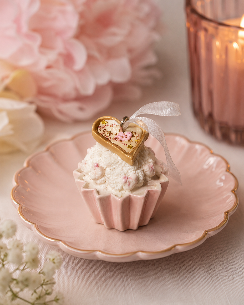 Pink and white cupcake ornament with a gold heart charm displayed on a pink scalloped plate beside a candle and pink flowers