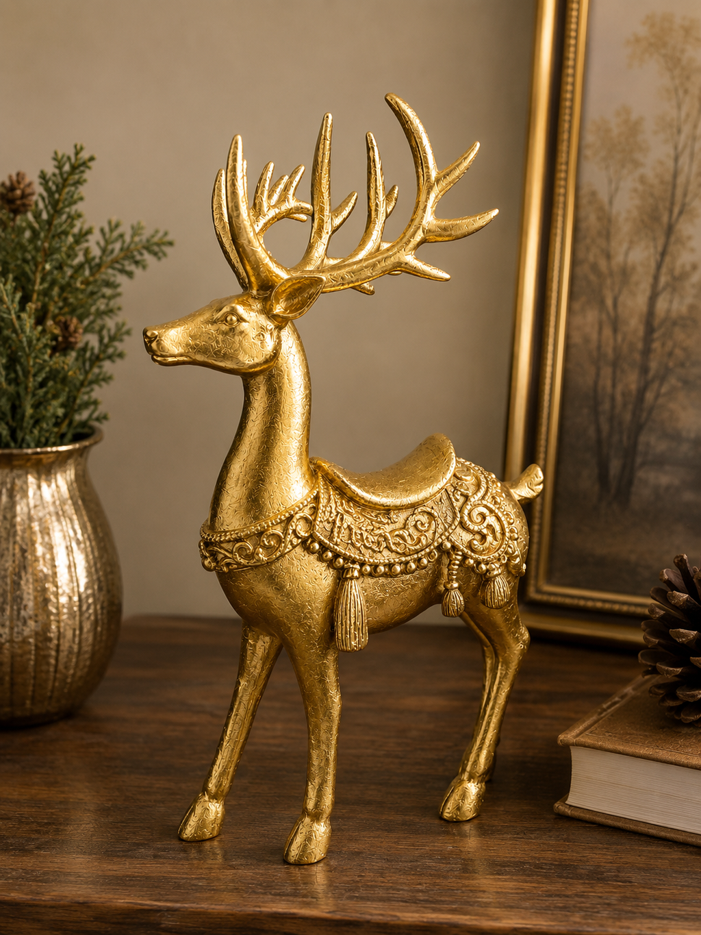 Gold resin standing reindeer figurine with ornate saddle, scrollwork and tassel detail, styled on a timber surface with gold vase, greenery and framed artwork