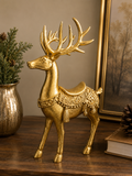 Gold resin standing reindeer figurine with ornate saddle, scrollwork and tassel detail, styled on a timber surface with gold vase, greenery and framed artwork