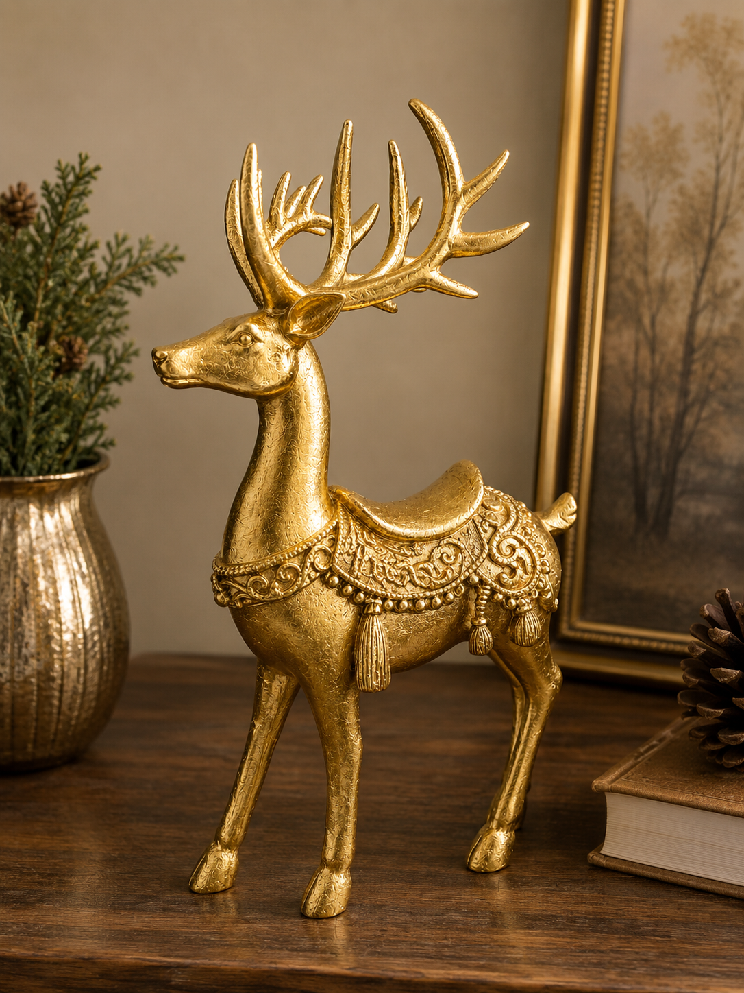 Gold resin standing reindeer figurine with ornate saddle, scrollwork and tassel detail, styled on a timber surface with gold vase, greenery and framed artwork