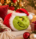 Santa Grinch Spudster plush resting on a cream knit throw surrounded by Christmas ornaments, fairy lights, red cushions, and a striped gift box — festive Christmas home setting