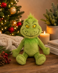 The Grinch plush soft toy sitting on a wooden surface styled with a lit Christmas tree, cream candle, pinecones, and red berries in the background — cosy festive setting