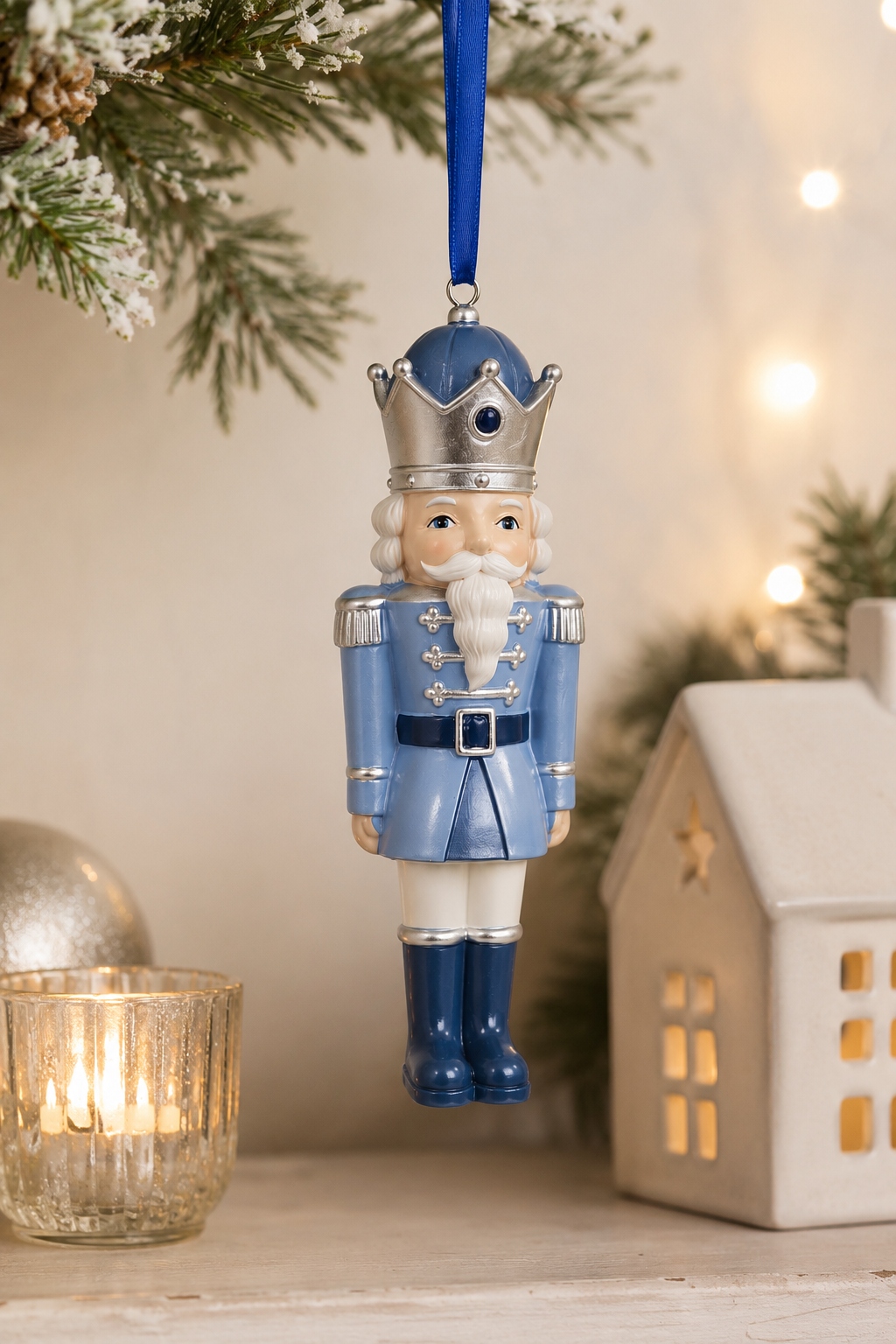 Blue and silver nutcracker king Christmas ornament hanging from a frosted pine branch, styled with a glowing mercury glass candle holder and ceramic lit house, wearing a silver crown and powder blue military jacket with white beard