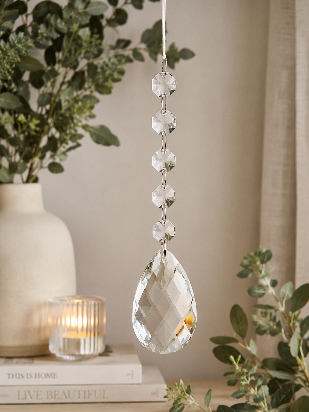 Clear crystal pear drop ornament hanging in a neutral home interior styled with eucalyptus, stacked books, and a lit ribbed glass candle holder, showing the elegant chandelier-style design