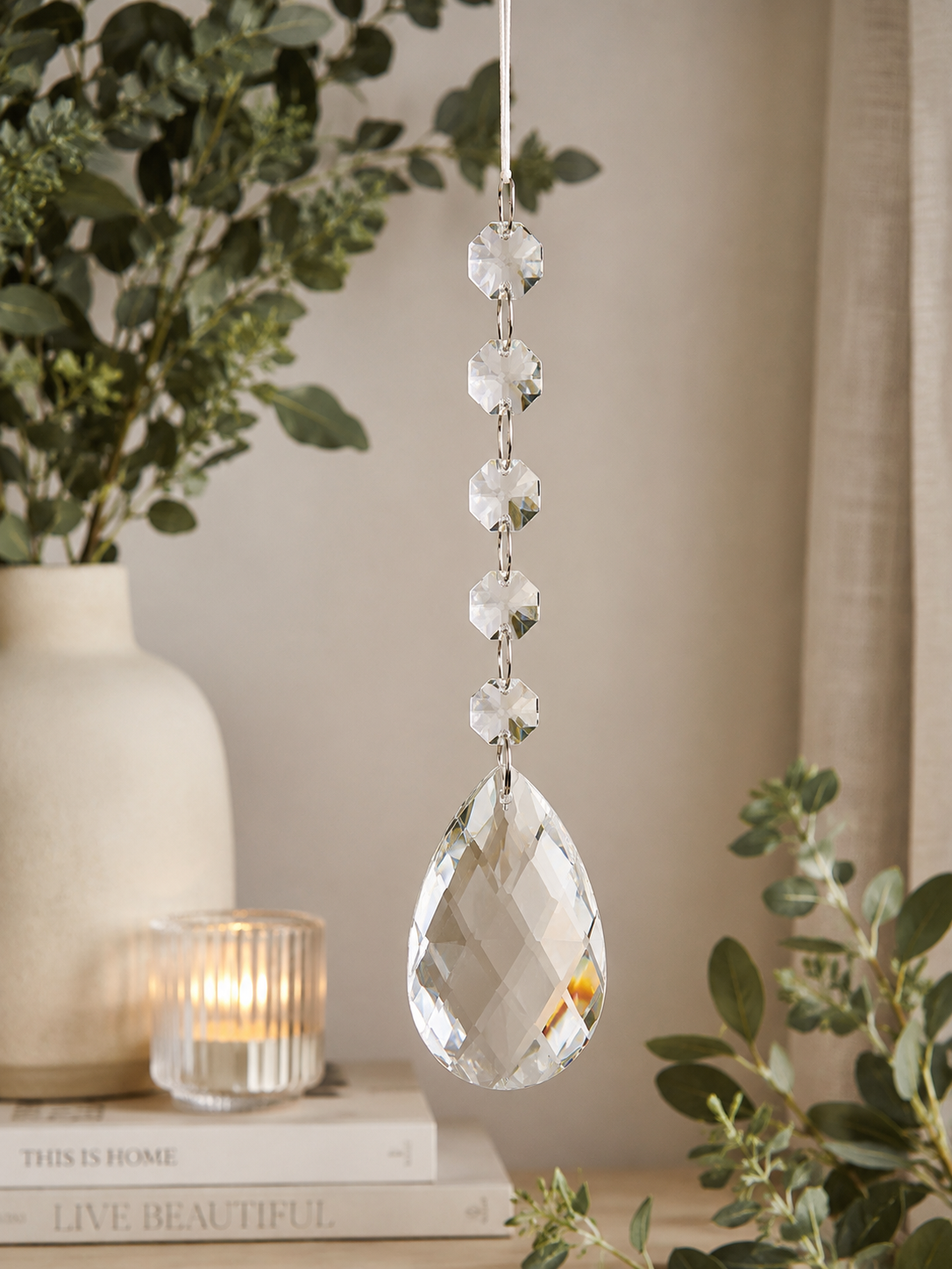 Clear crystal pear drop ornament hanging in a neutral home interior styled with eucalyptus, stacked books, and a lit ribbed glass candle holder, showing the elegant chandelier-style design