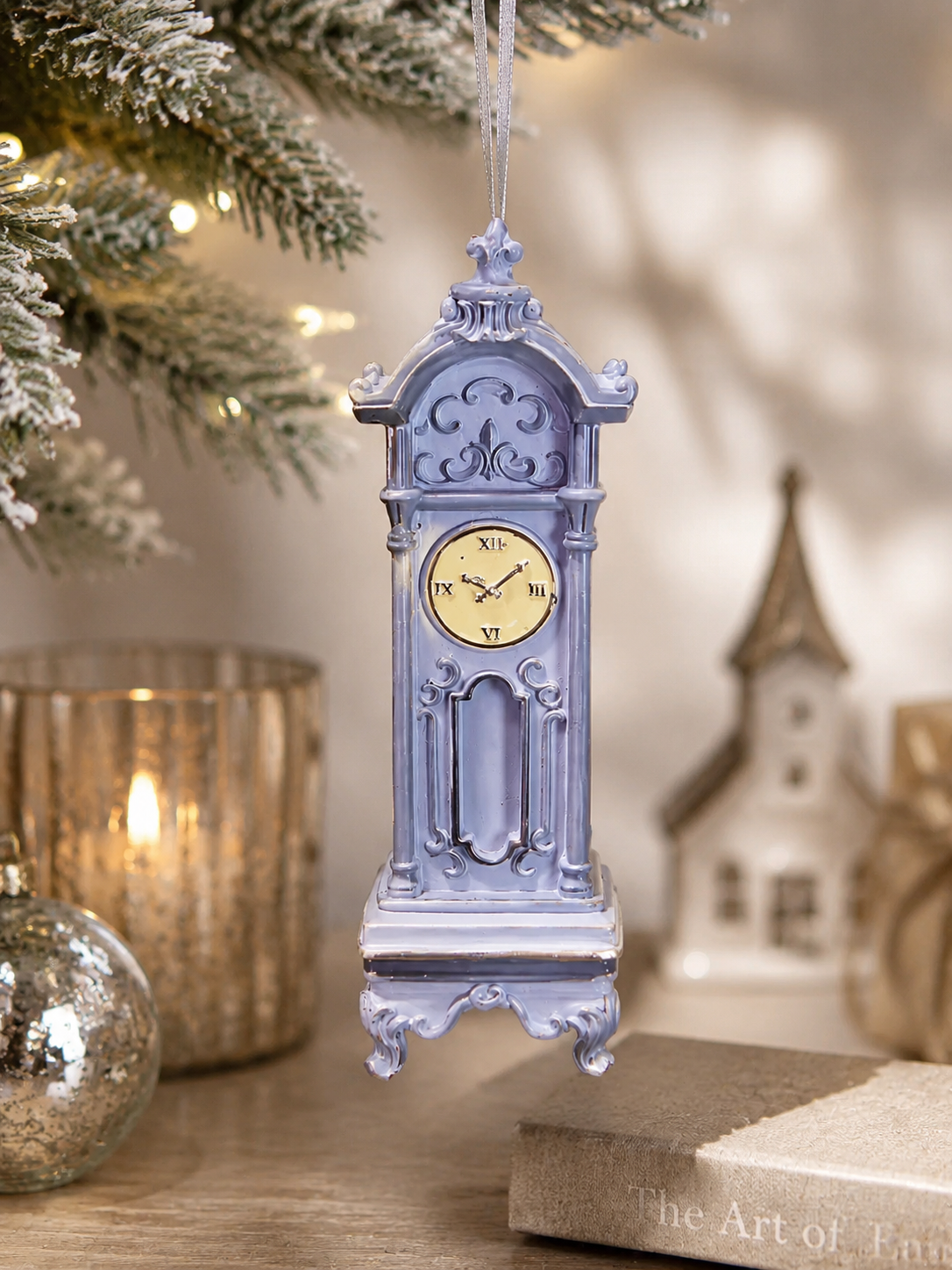Dusty blue grandfather clock Christmas ornament hanging from a frosted Christmas tree, styled with gold mercury glass candle holders, silver baubles and a miniature house figurine