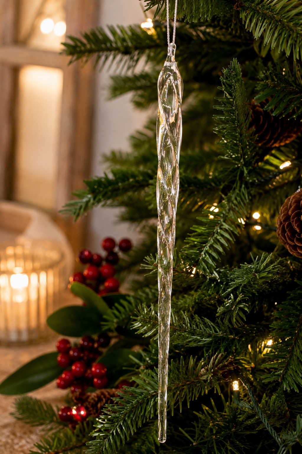 Clear glass icicle ornament on a lit Christmas tree with warm candlelight, red berry stems and pine cones in the background, glowing in the warm light