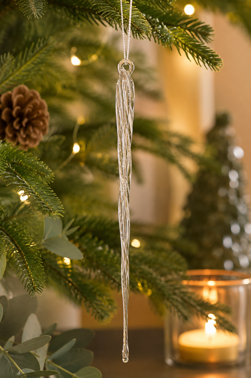Clear glass braided icicle ornament glowing on a lit Christmas tree in warm candlelight, with eucalyptus and a glass tea light holder in the background, highlighting the intricate twisted texture