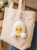 Bobby Egg plush bag charm keyring clipped to a cream canvas Assembly Label tote bag, worn over the shoulder against a warm neutral background