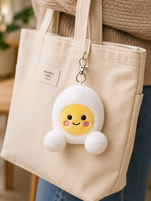 Bobby Egg plush bag charm keyring clipped to a cream canvas Assembly Label tote bag, worn over the shoulder against a warm neutral background