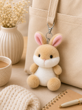 Ella Bunny plush bag charm keyring leaning against a cream canvas tote bag on a neutral surface, styled with a ceramic coffee cup, white ribbed vase with dried flowers, spiral notebook, pen, and knit fabric