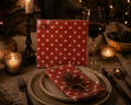 Pack of red Gala paper napkins with white star print propped on a candlelit Christmas dinner table with a folded napkin tied with twine and pine sprig on a ceramic plate, wine glass and Christmas tree in the background — Mistletoe Home